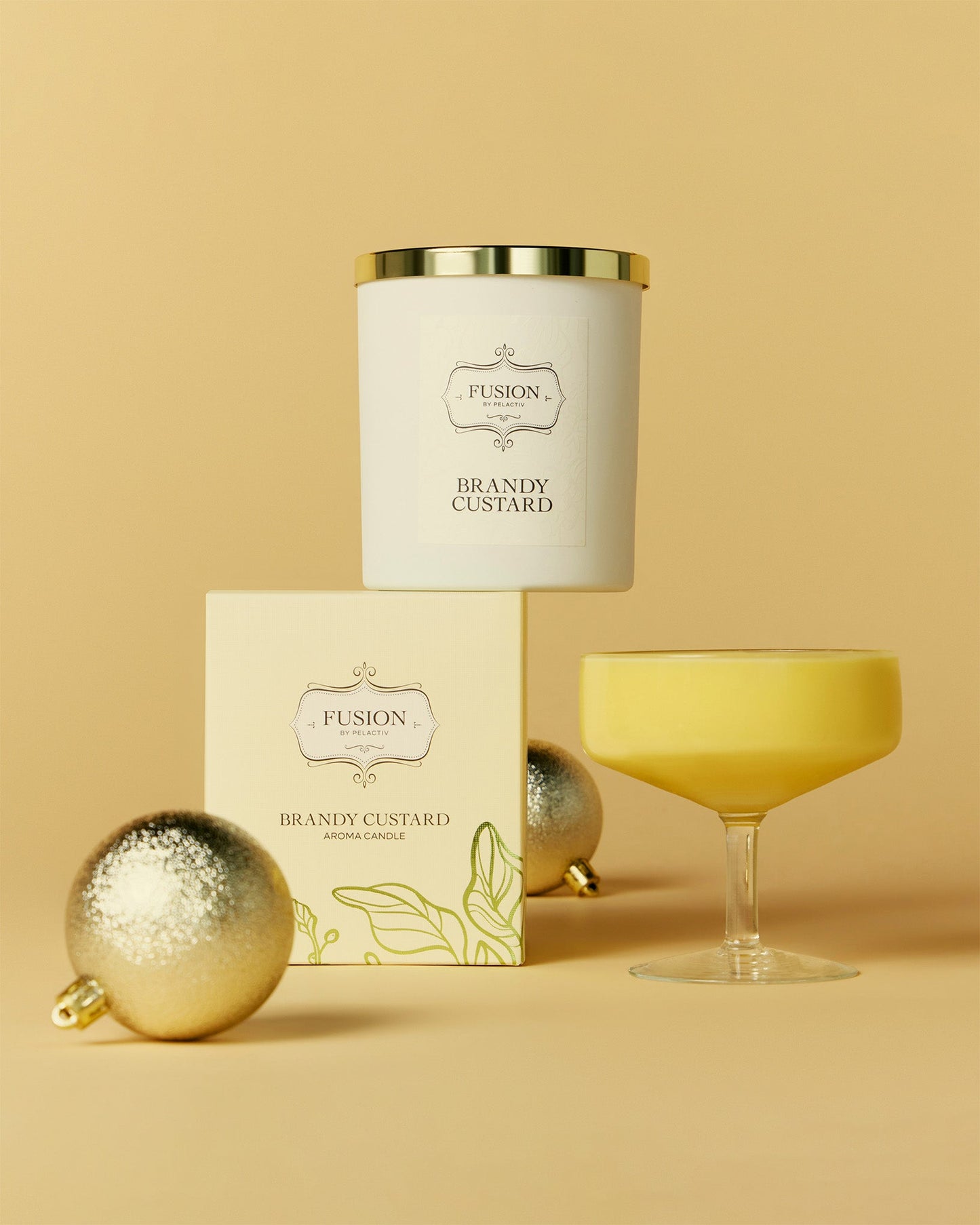 A decorative scene with a candle labelled Brandy Custard, glass with custard in it, and the packaging box against a vibrant yellow backdrop, featuring decorative Christmas Ornaments.