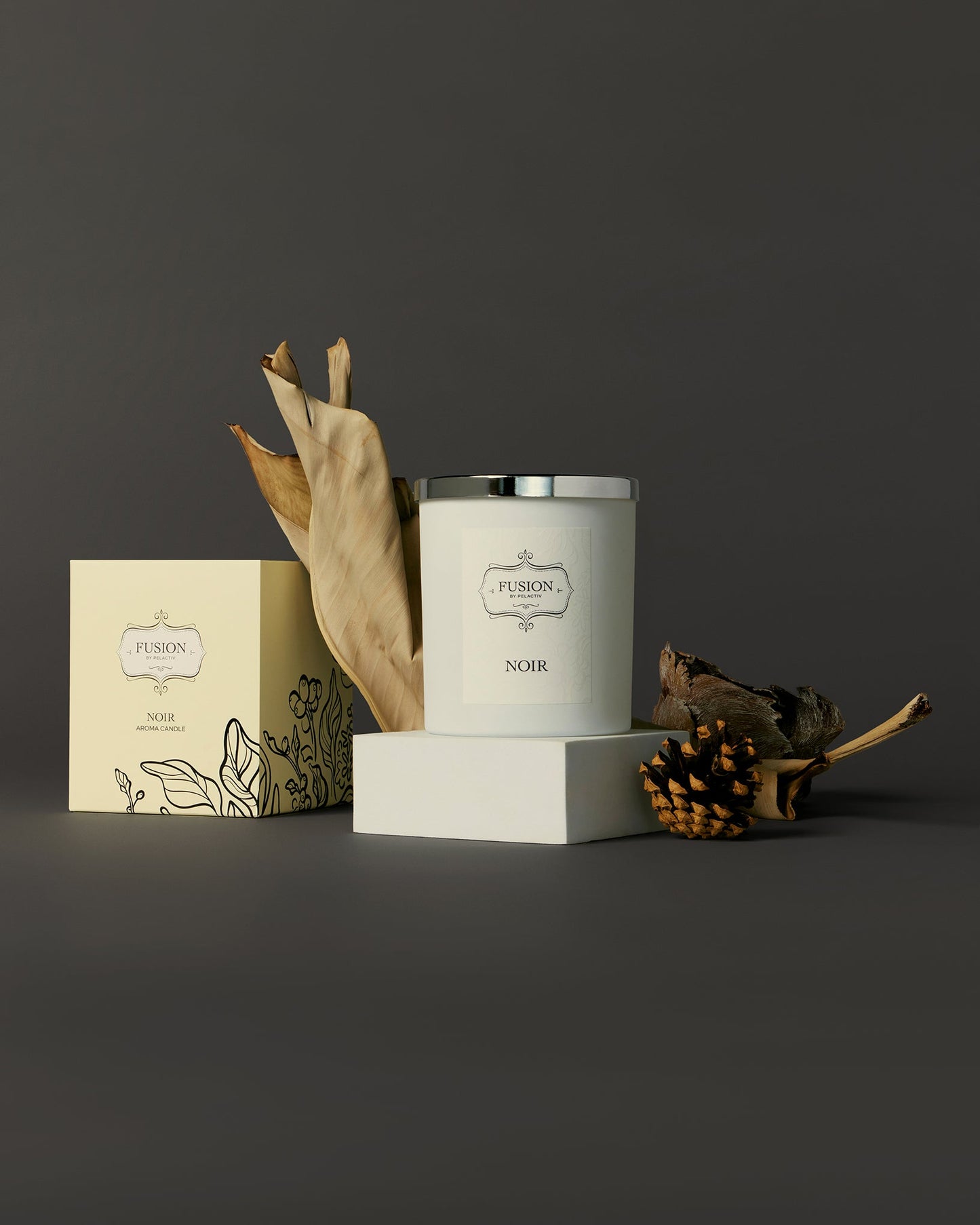 Elegant candle display featuring a white candle with a silver lid beside its box, accented by dried leaves and pinecones, set against a dark backdrop.