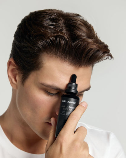Man holding a black dropper bottle labeled 'Mr. Jones Hair Density Serum' close to his face against a plain background