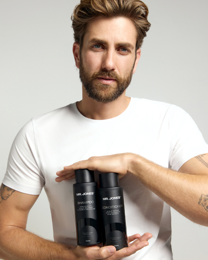 Man holding two black bottles labeled 'Mr. Jones Shampoo and Conditioner' against a light grey background